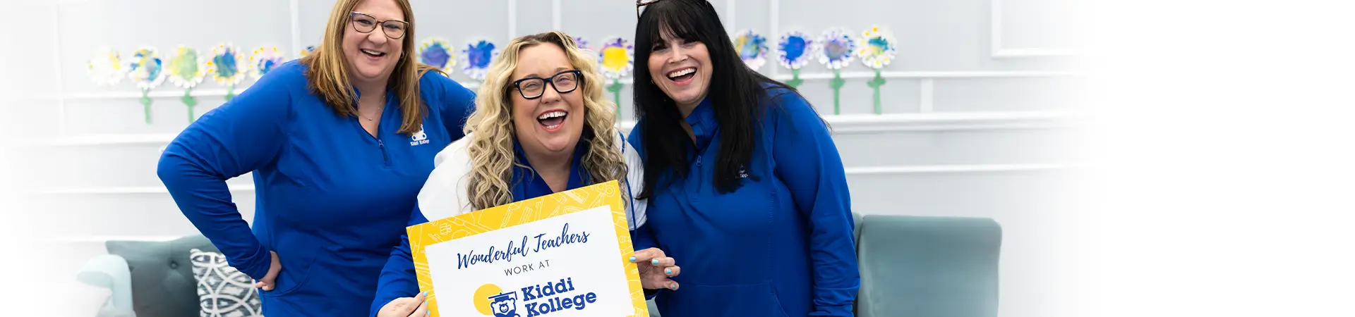 Teachers Smiling holding Work At Kiddi Kollege sign